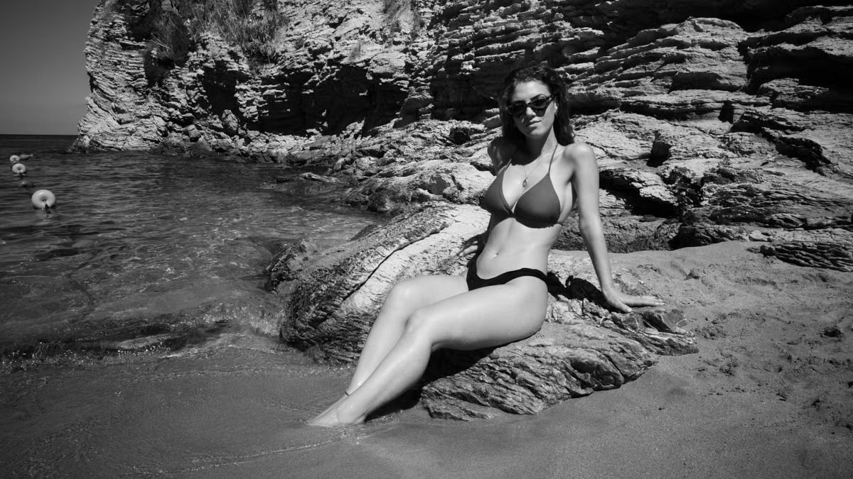 A woman in sunglasses poses on a rocky beach, enjoying a sunny day by the sea.